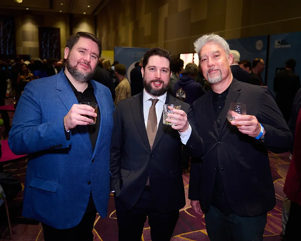 Three men dressed in suits attending a social event, holding drinks and posing for the camera in a conference or banquet hall.