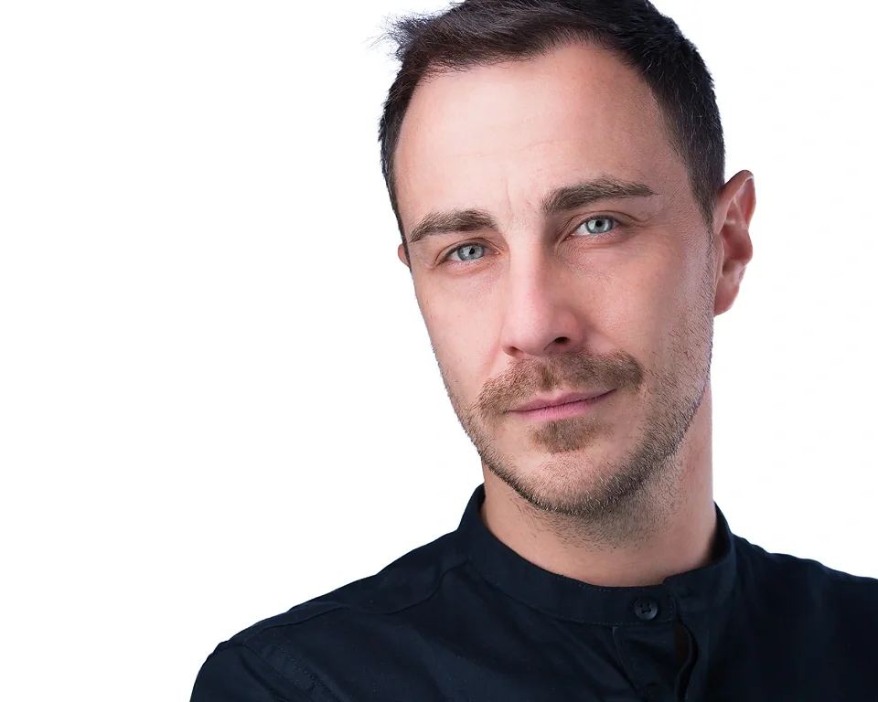 A man with short dark hair, blue eyes, and a light beard and mustache looking at the camera with a slight smile, wearing a dark high-collared shirt, against a plain white background.