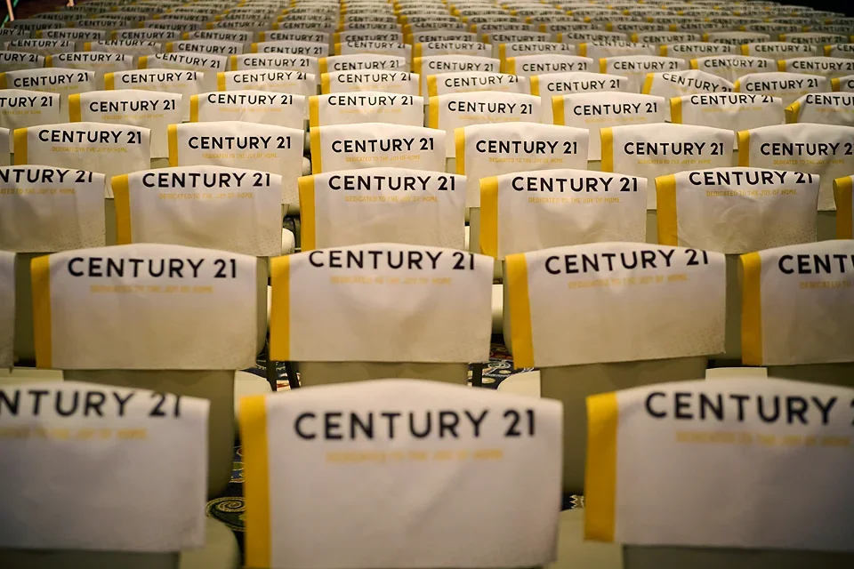 Rows of chairs with white covers and yellow accents, printed with 'CENTURY 21' in black, arranged in a conference or event hall.