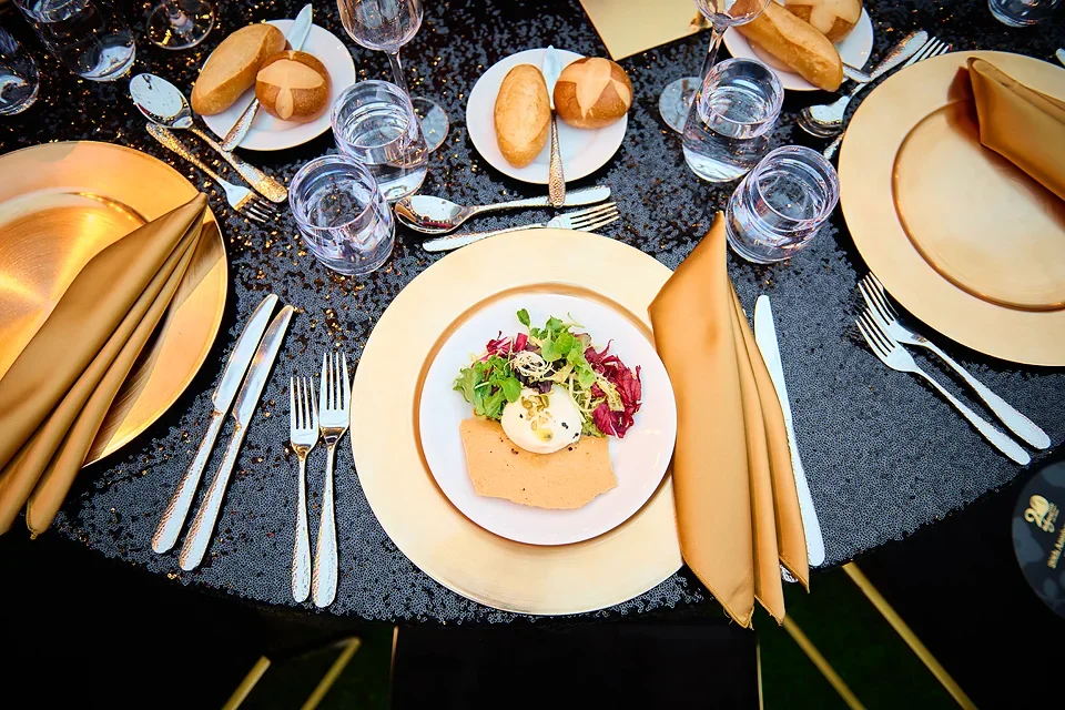 Elegant formal dining table setup with gold plated cutlery, folded gold napkins, water glasses, bread rolls on small plates, and a plated appetizer with greens and a small cup of sauce.