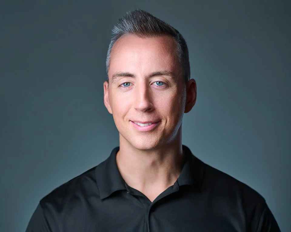 A young man with short dark blonde hair, blue eyes, and light skin, wearing a black collared shirt, smiling softly, against a neutral gradient background.