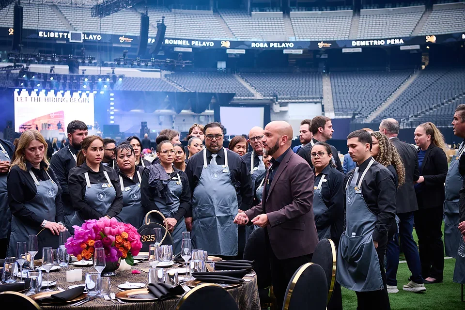 People gathered around a table wearing aprons, listening to a speaker in a stadium setting, with a floral centerpiece and set dining table in the foreground.