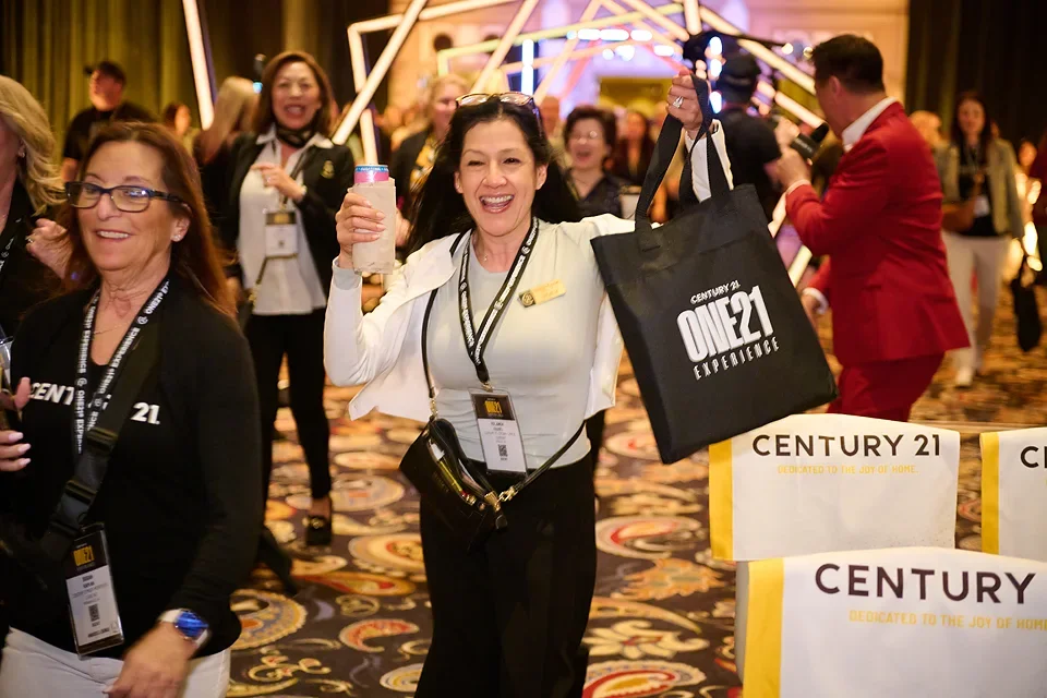 A group of smiling women at a conference or event, with one woman holding a Century 21 bag and a drink, walking on a colorful patterned carpet.