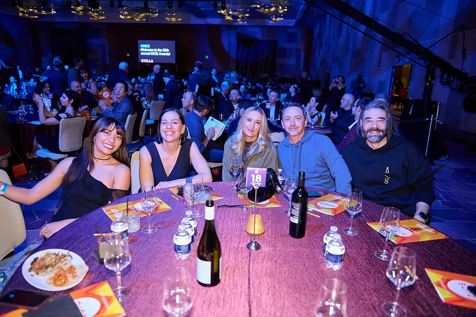 Five people sitting at a round dinner table during an event, with other guests in the background, and a screen on stage displaying 'GREO Welcome to the 2023 Annual DICE Awards Stella'.