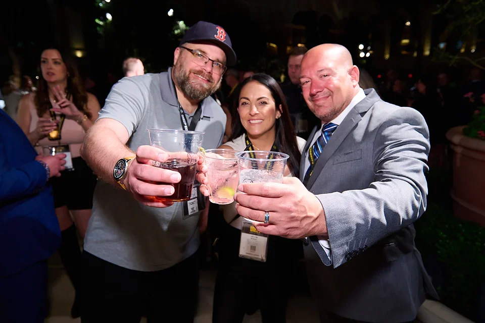 Three people smiling and holding drinks at a social event, with a woman and two men.