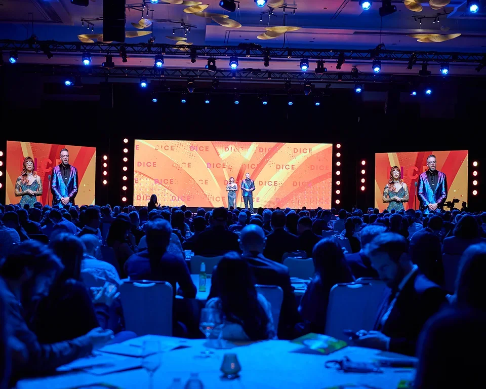 An audience watching a presentation on stage at a conference or event, with three large screens displaying two presenters.