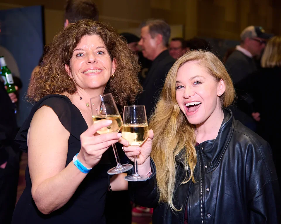 Two women at a social event holding glasses of white wine and smiling at the camera, with a crowded background.