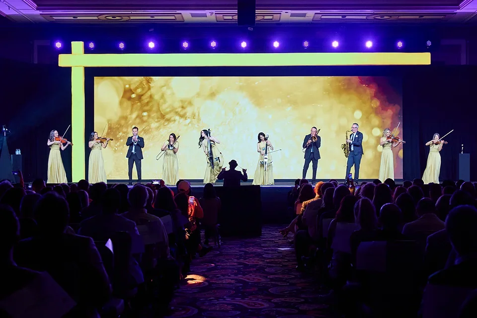 Orchestra performing on stage with a conductor in front, featuring string instruments and a large digital backdrop, in front of an audience in a dimly lit venue.