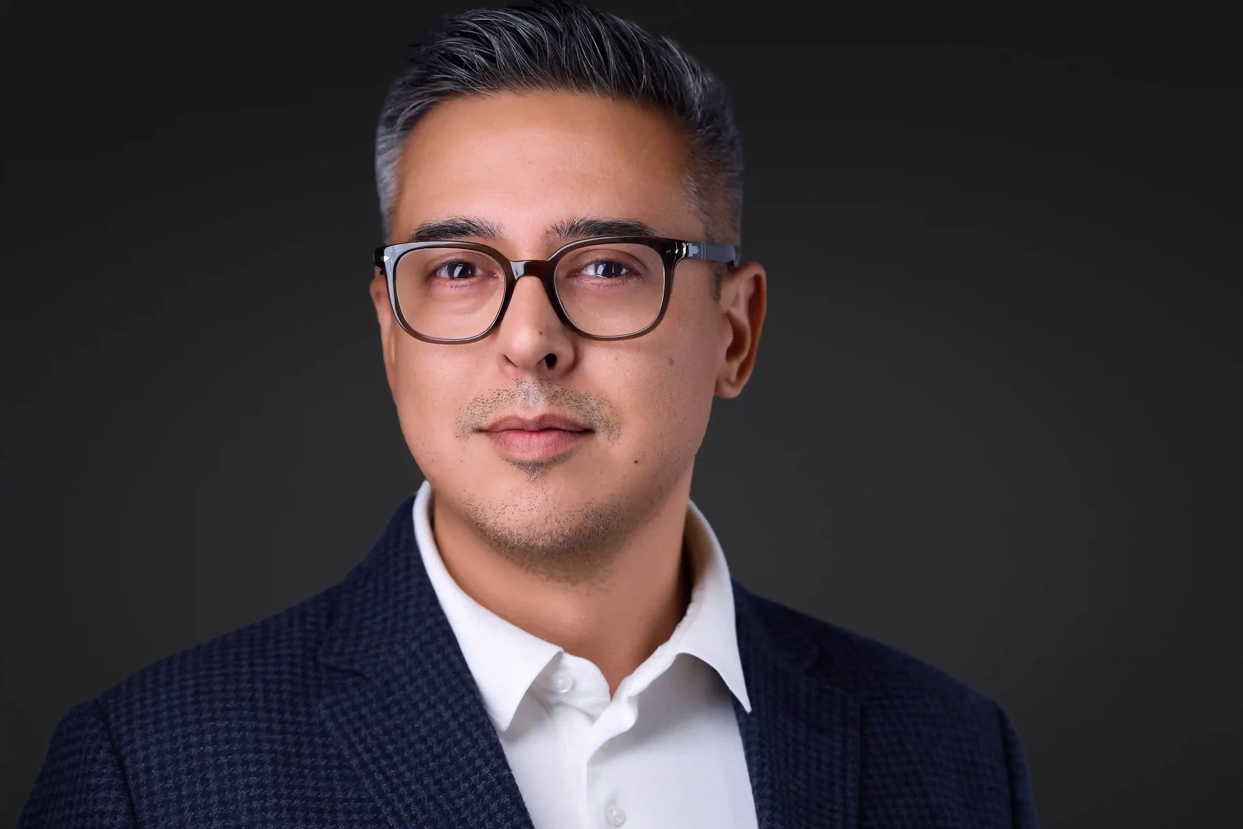 Professional headshot of a man with short dark hair wearing glasses and a navy blazer with a white shirt, against a dark background, photographed at Pandorica Headshot Studio in Las Vegas