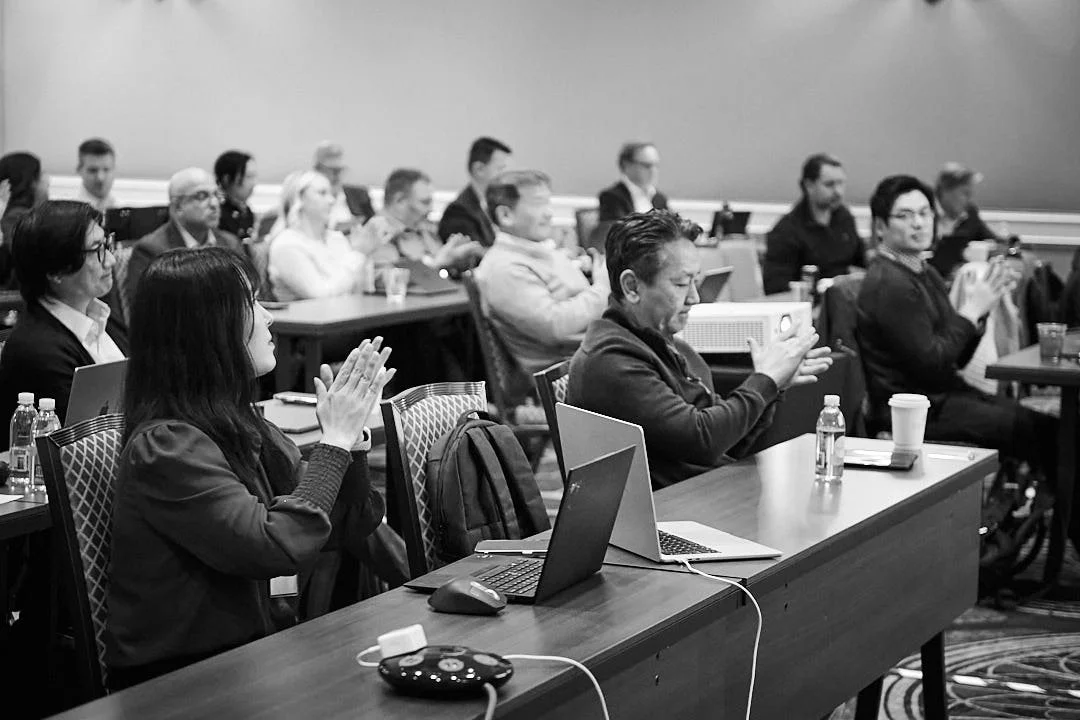 People sitting at tables in a conference room, some clapping and others taking photos, with laptops and water bottles on the tables.