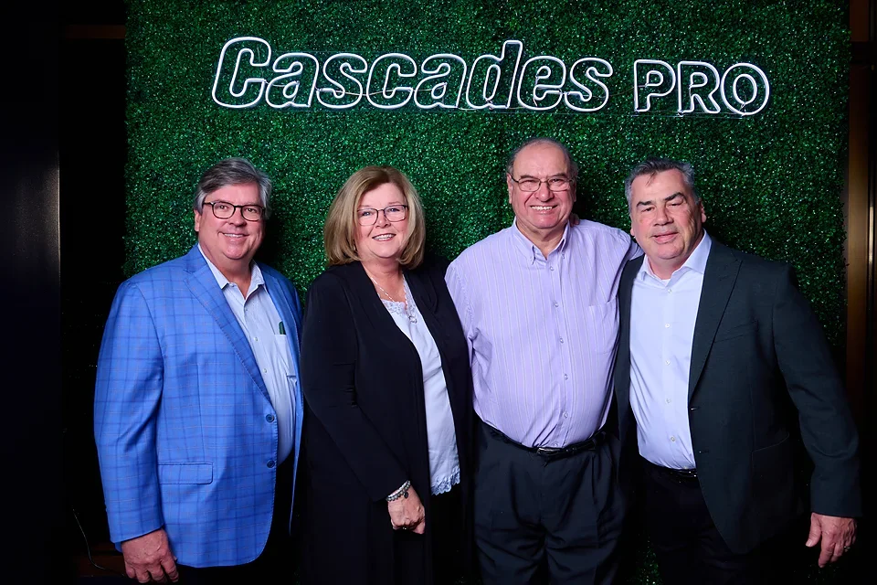 Four business professionals, two men and two women, dressed in business attire, standing together in front of a green textured wall with 'Cascades PRO' illuminated sign.