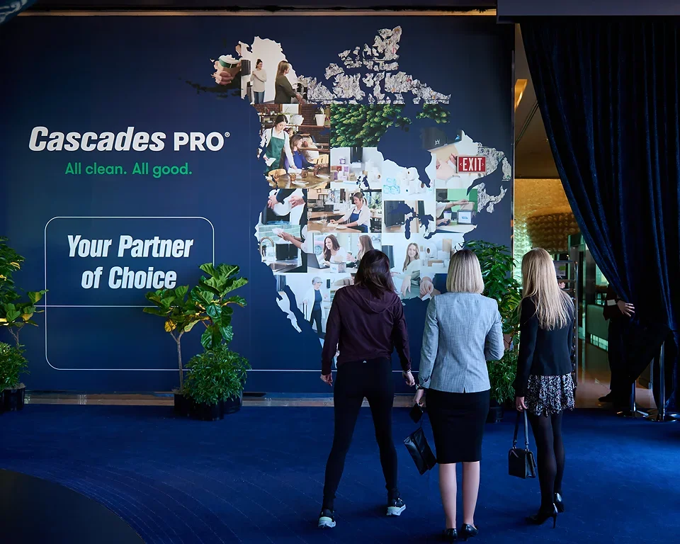 Three women walking towards a large wall display about Cascades PRO, with a map of Canada made up of photos, and indoor plants on either side.