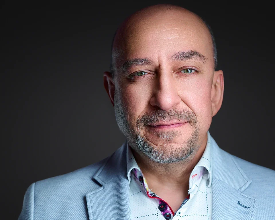 A middle-aged man with a goatee and blue eyes, wearing a light gray blazer over a patterned shirt, smiling gently against a dark background.