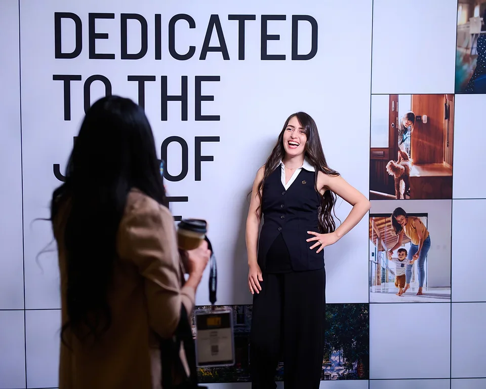 A woman with long dark hair smiling and posing for a photo in front of a large sign that reads 'Dedicated to the' with the rest of the text obscured, while another woman with long dark hair holding a camera takes her picture.