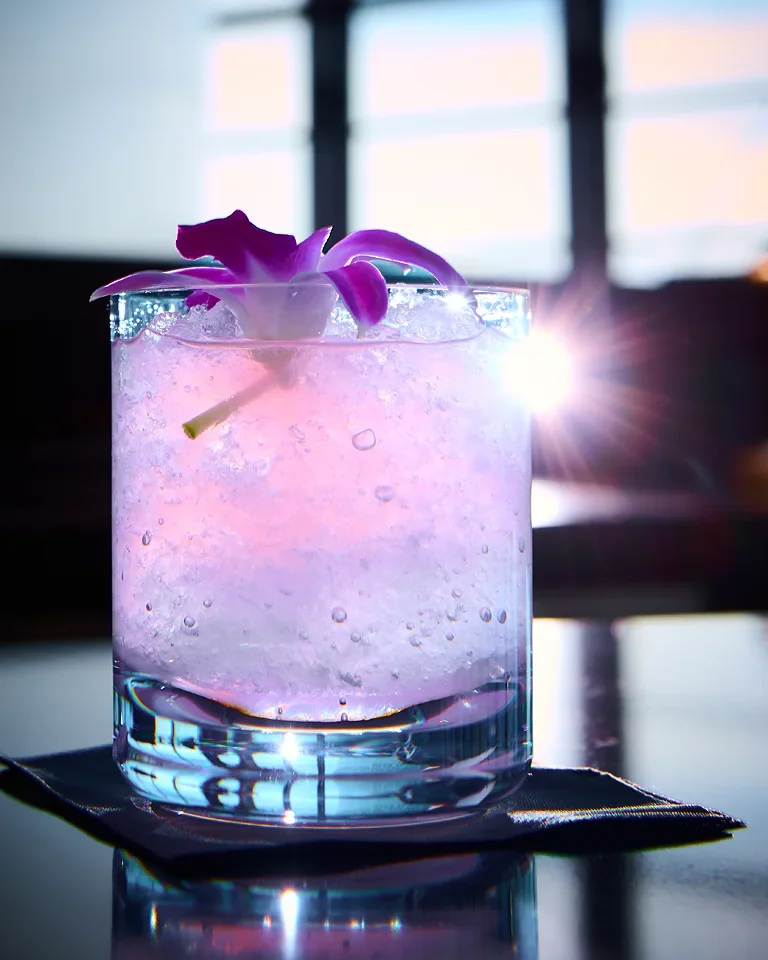 A pink and purple cocktail with an orchid flower garnish in a clear glass, with sunlight and window in the background.