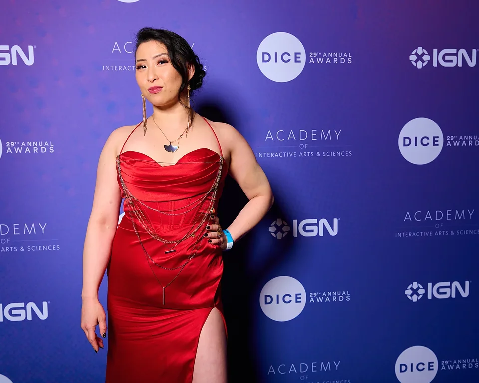 Woman in a red gown with jewelry, posing at the DICE 29th Annual Awards event with a blue backdrop featuring logos and text.