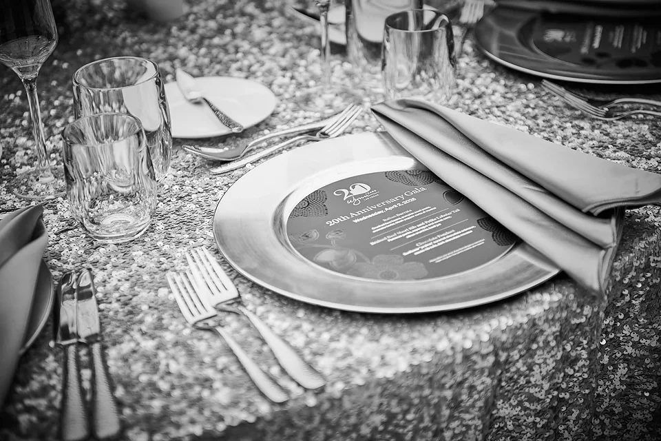 Table setting with a concert-style plate, wine glasses, water glasses, napkins, silverware, and a glittery tablecloth, with some plates and glasses in the background.