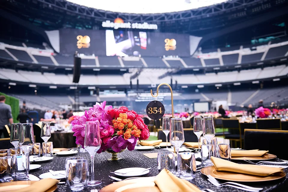 Formal event setup inside a large stadium with a floral centerpiece, glasses, and tableware, with a digital screen displaying the number 20 in the background.
