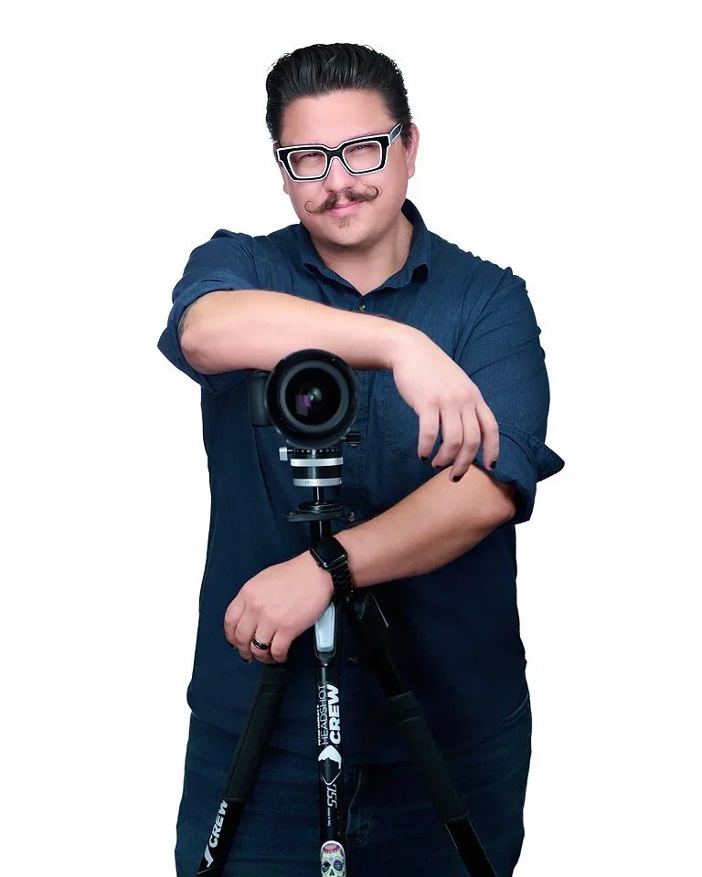A man with glasses, a mustache, and a goatee, wearing a dark blue shirt, taking a selfie in front of a camera mounted on a tripod against a plain white background.