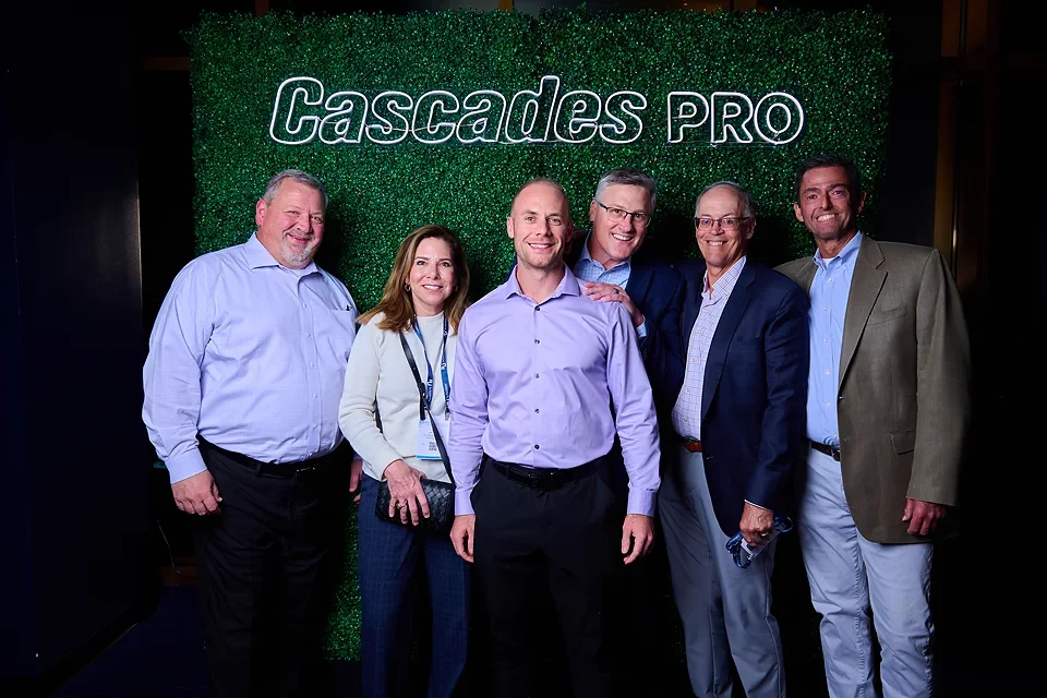 Six people, three men and three women, standing together in front of a green backdrop with the words 'Cascades PRO' displayed. They are dressed in business casual attire and are smiling.
