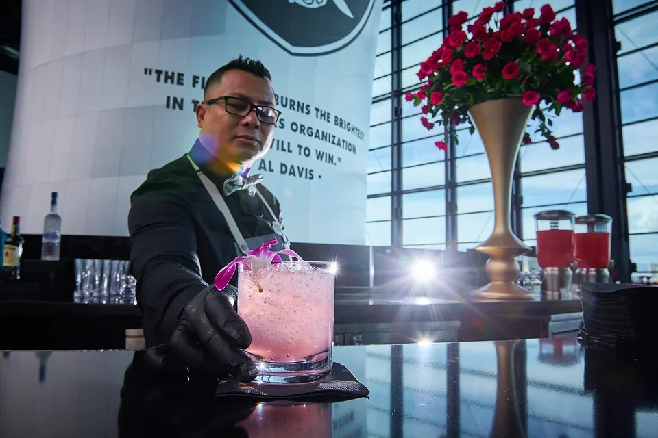 A bartender in black attire and gloves presenting a pink cocktail with orchids garnish at a bar with large windows and a flower arrangement in a gold vase in the background.