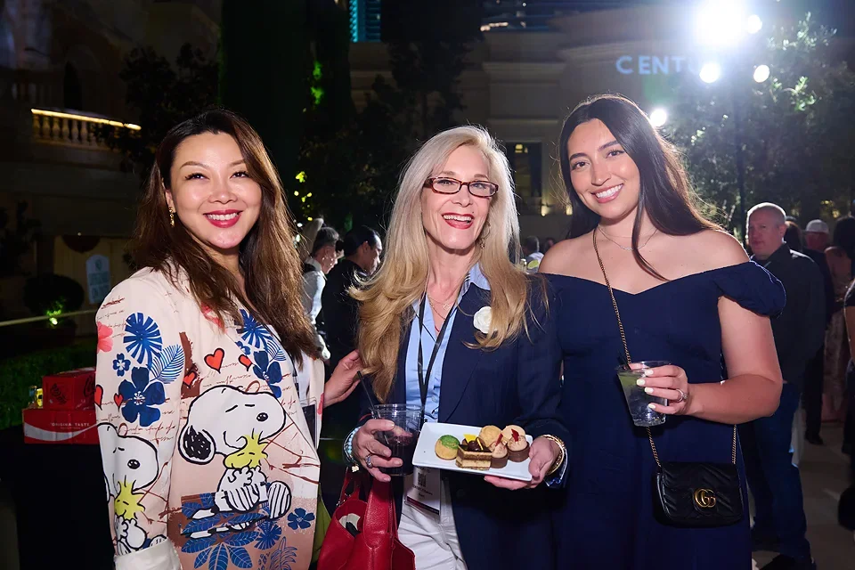Three women smiling at a social event, two holding drinks and one holding a plate with desserts, outdoors at night with others in the background.
