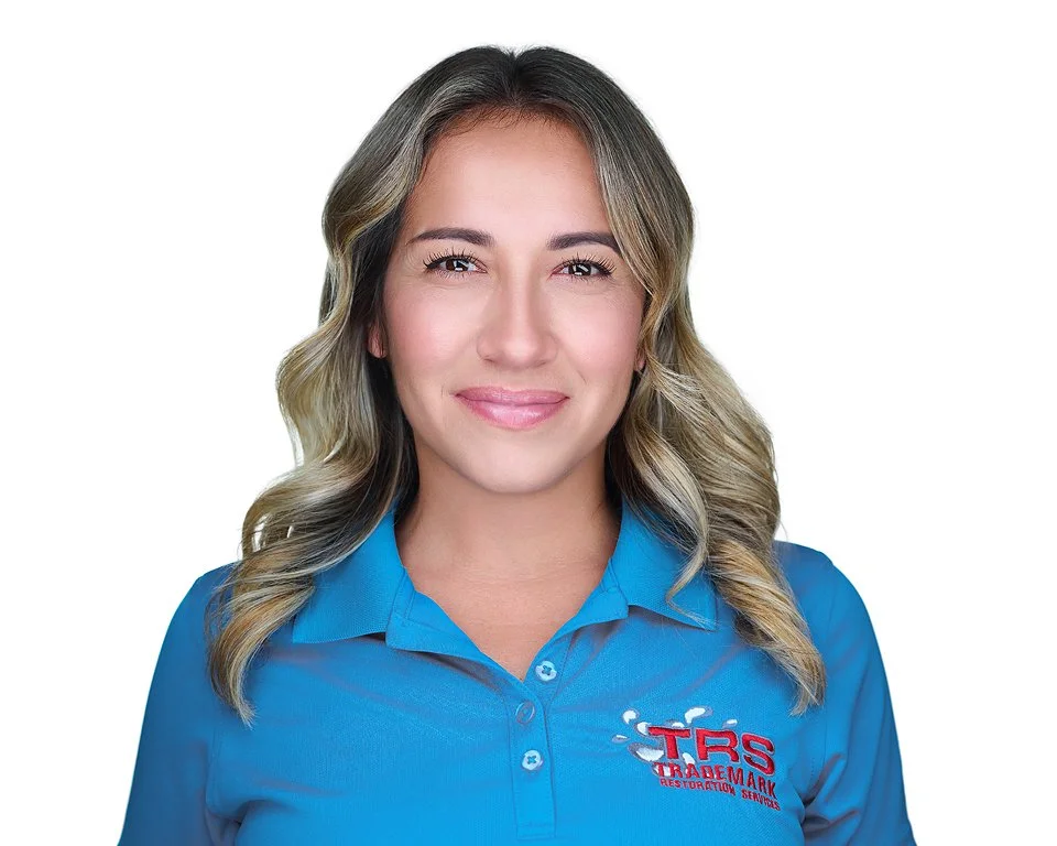 A woman with wavy blonde hair, wearing a blue polo shirt with a logo for TRS TradeMark Restoration Services, smiling at the camera.