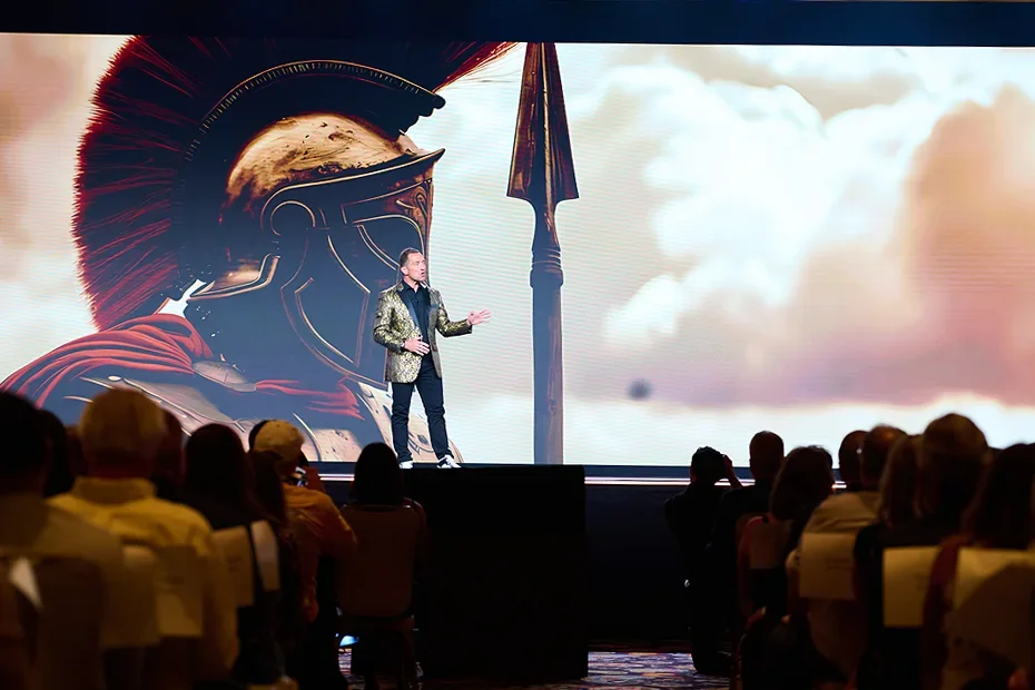 A man giving a presentation on stage in front of a large screen displaying an ancient Roman soldier with a helmet and spear, with an audience seated and listening.
