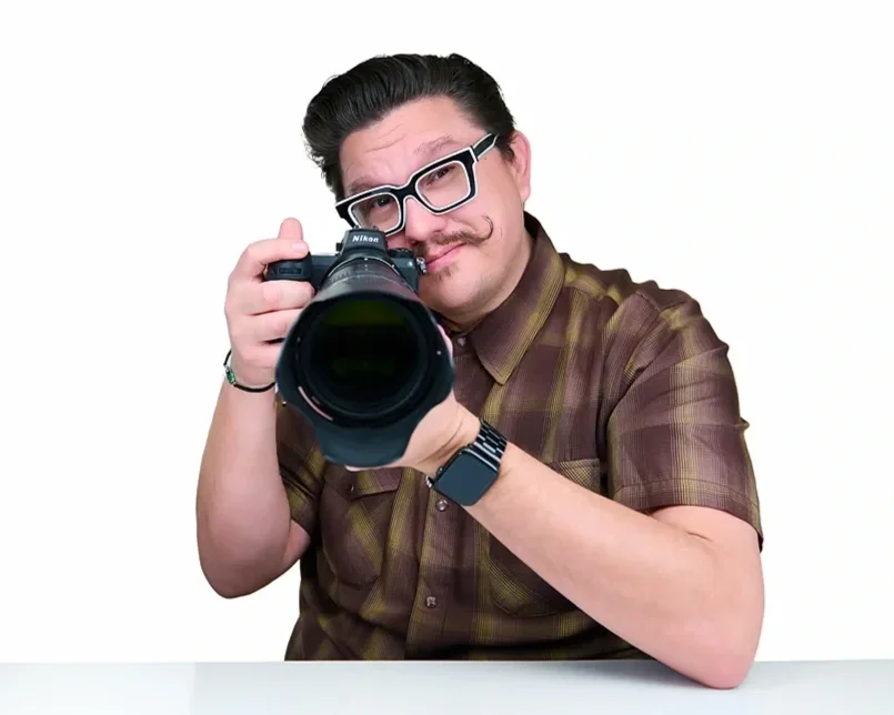 Mondo Rojas, glasses, and a mustache taking a photo with a Nikon camera, wearing a brown plaid shirt and a watch, against a plain white background. Pandorica headshot studio