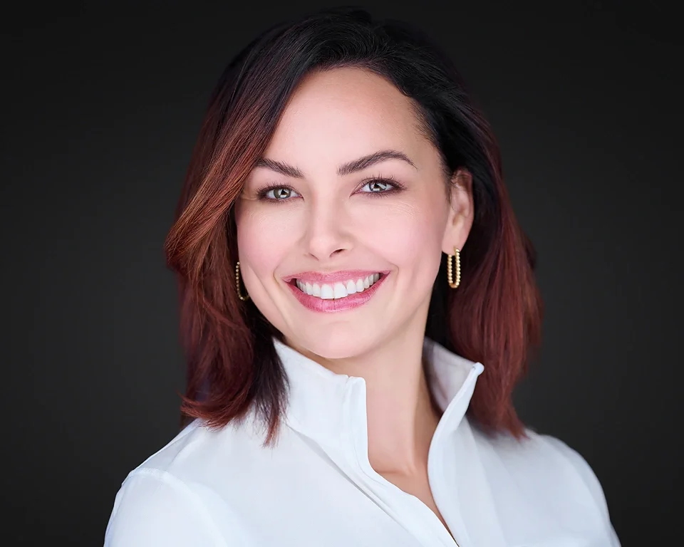 Portrait of a woman with shoulder-length hair, smiling, wearing a white collared shirt and gold hoop earrings.