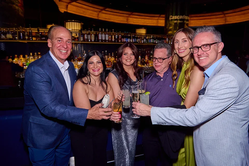 Group of six people celebrating with drinks at a bar or lounge, smiling for the camera.