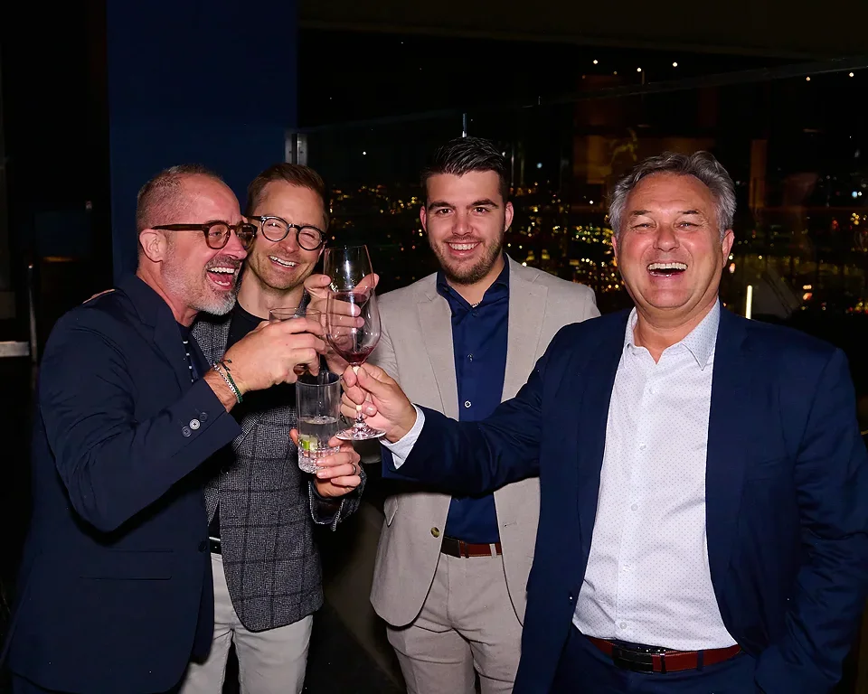 Five men in business attire celebrating with drinks at a dinner party, city lights visible through the window behind them.