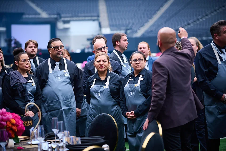 Group of people wearing aprons attending a cooking class or demonstration in an indoor stadium or arena.