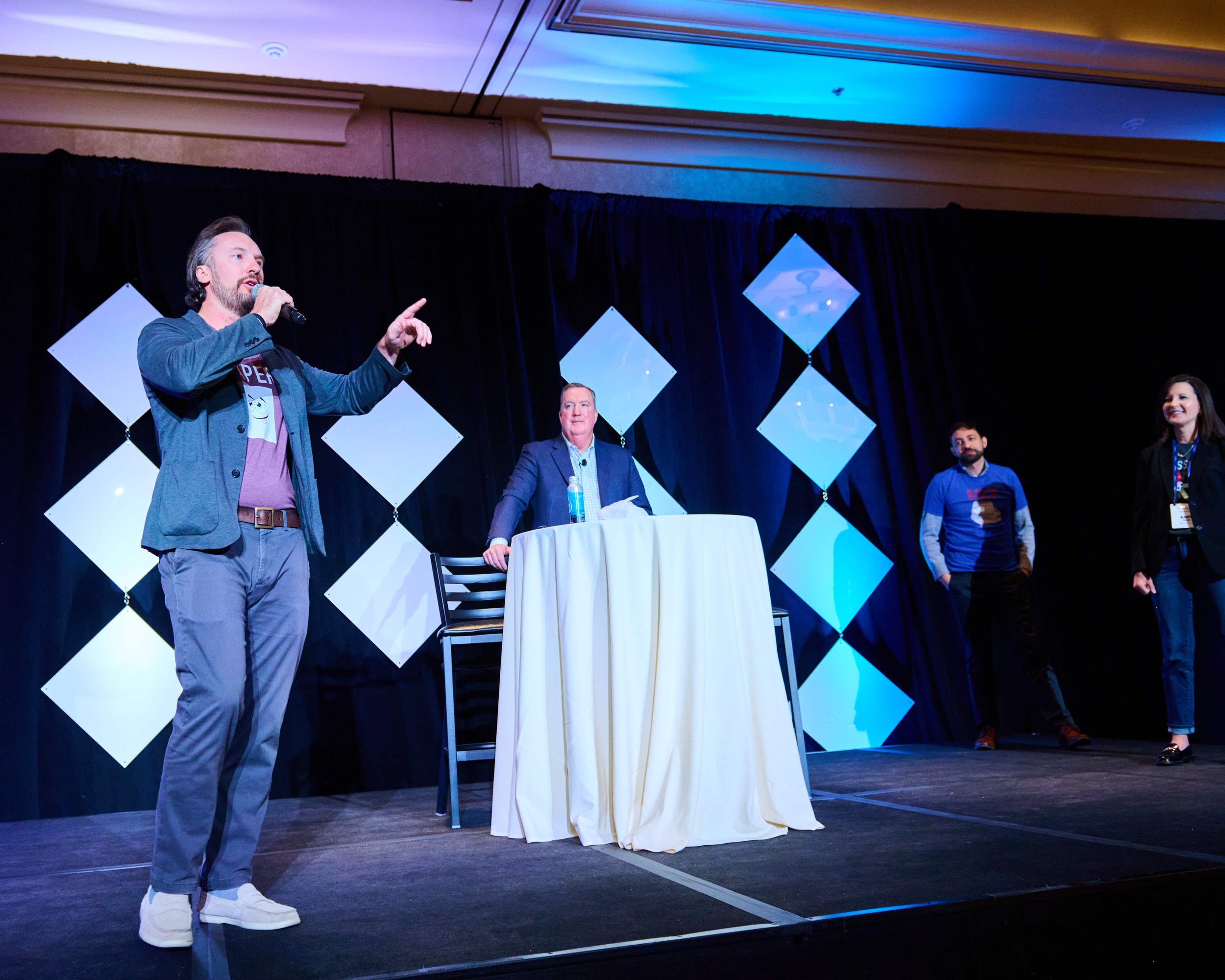 A man with a beard and long hair speaking into a microphone on stage, gesturing with his right hand. He wears a blazer, T-shirt, gray pants, and white shoes. Behind him, three people are smiling and listening. The stage has black curtains and a decor