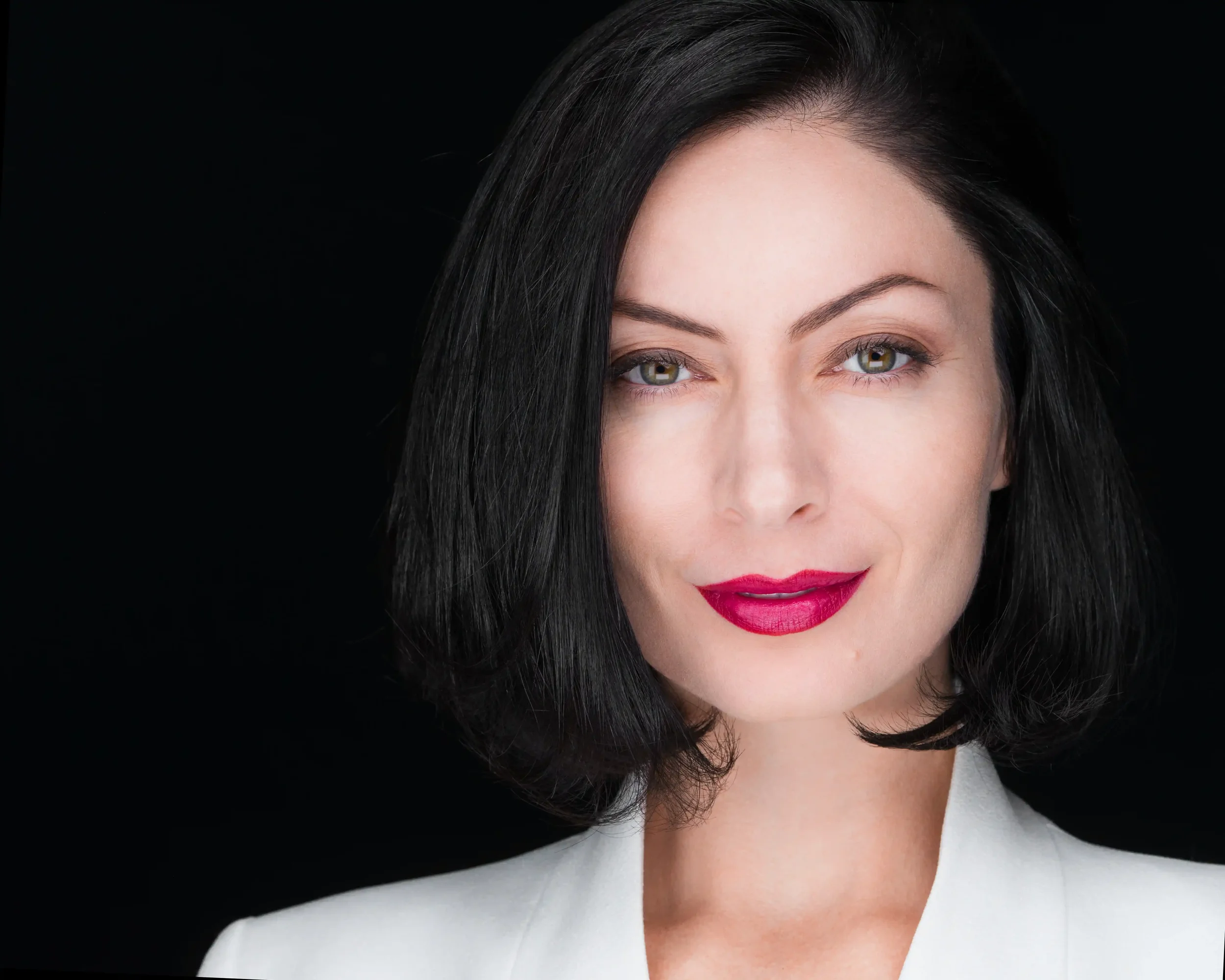 A woman with fair skin, black hair, and green eyes, wearing red lipstick and a white blazer, poses against a black background.