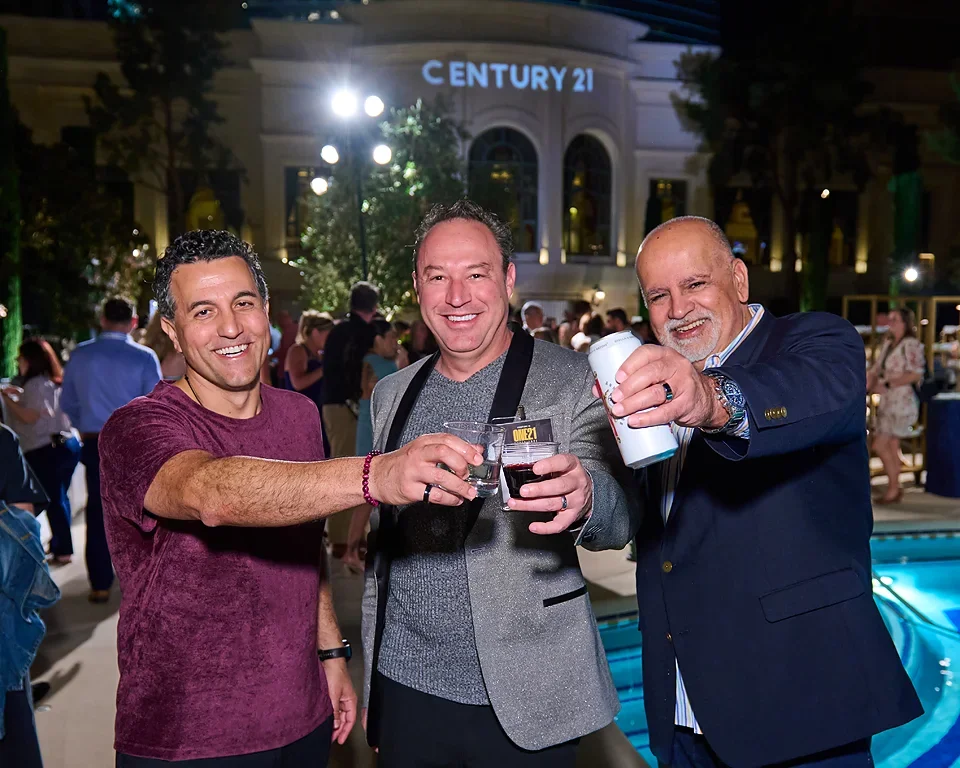 Three men smiling and raising drinks at an outdoor nighttime event, with a building labeled CENTURY 21 in the background and other people in the background.