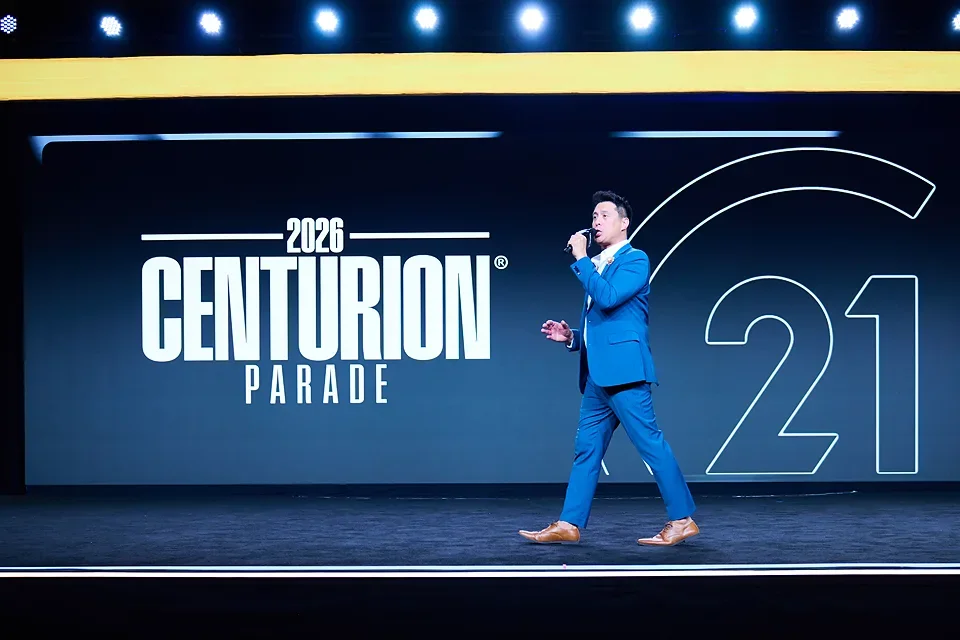 A man in a blue suit standing on stage with a microphone, presenting at the 2026 Centurion Parade event, with a large screen behind him displaying the event name and the number 21.