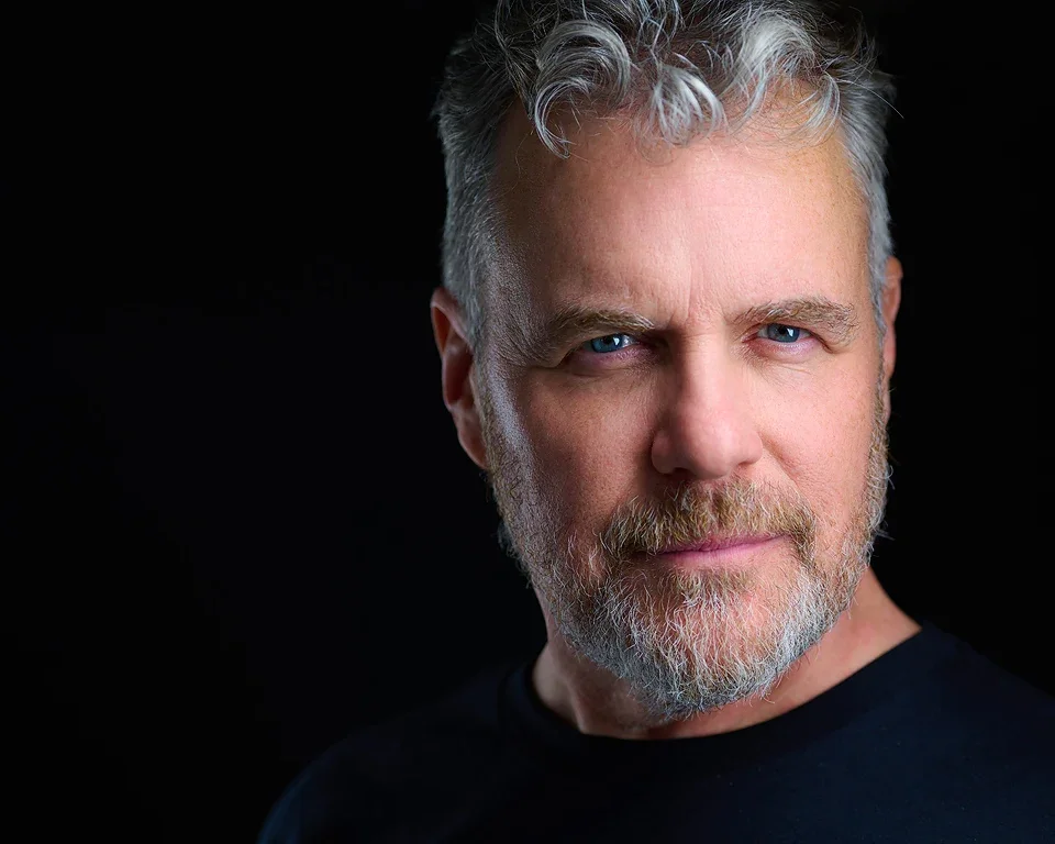 Close-up portrait of a middle-aged man with gray hair, blue eyes, and a beard, wearing a black shirt, against a black background.