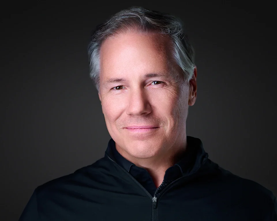 Headshot of a middle-aged man with gray hair, smiling, wearing a black zip-up jacket, against a dark background.