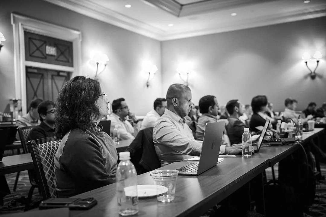 People attending a conference or seminar in a conference room, sitting at tables with laptops, bottled water, and notepads.