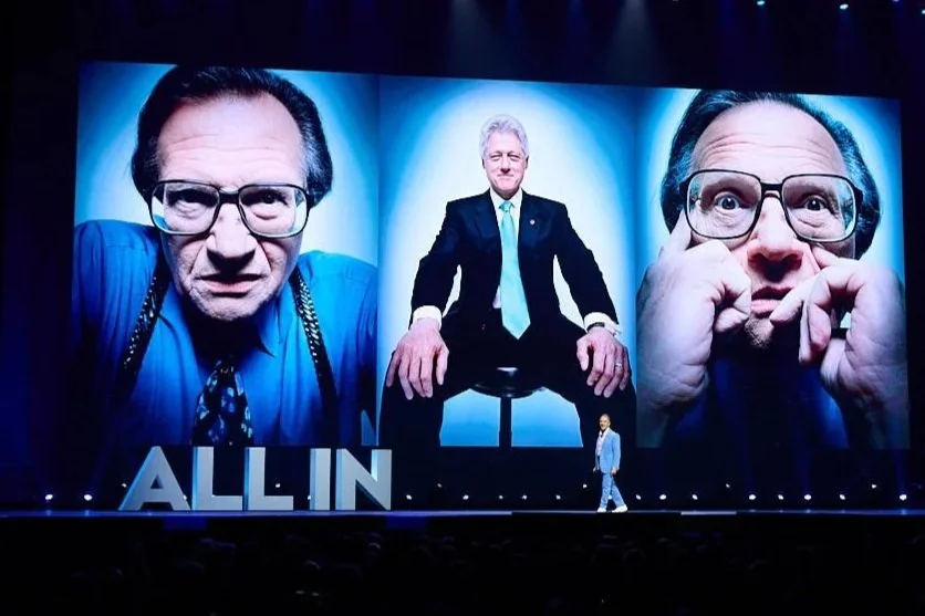 Stage with three large screens showing images of a man with glasses in various poses, and the words "ALL IN" at the bottom. A person stands on stage in front of the screens.
