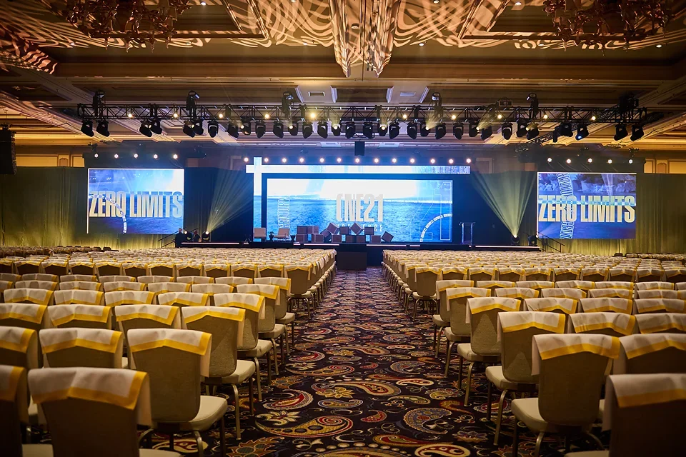 Large conference hall with rows of chairs facing a stage with big screens displaying 'ZERO LIMITS' and 'ONE21'.