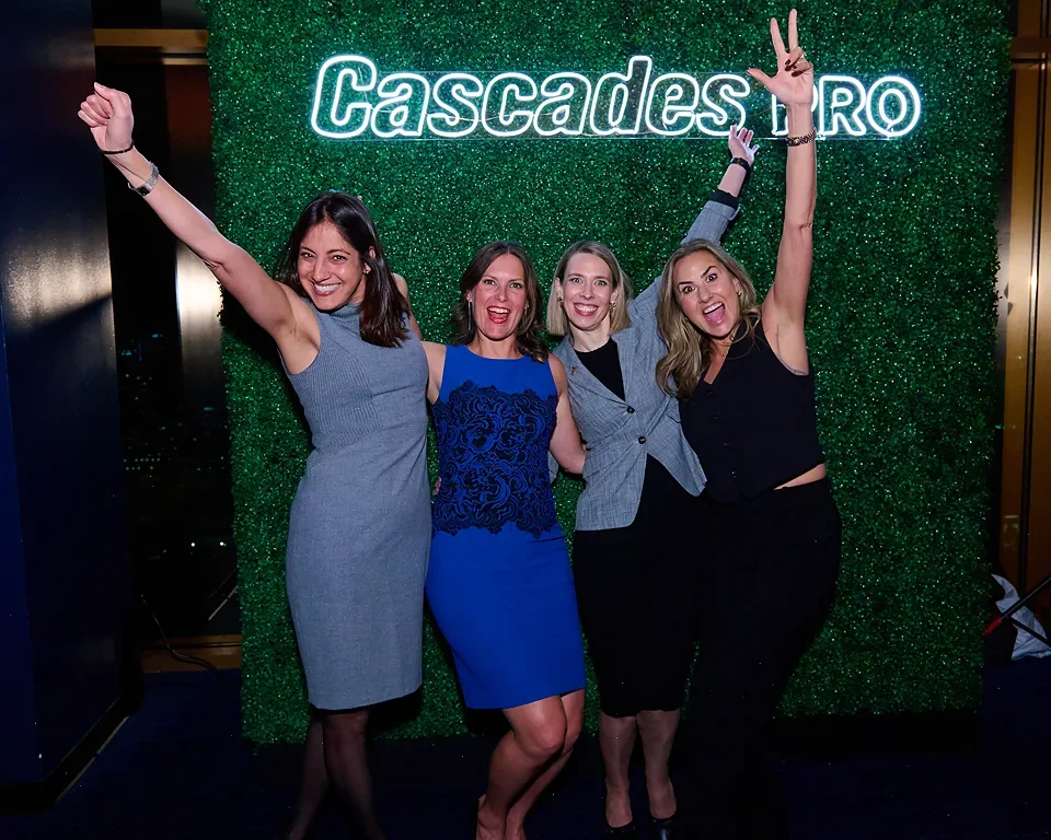 Four women posing in front of a green textured backdrop with 'Cascades' neon sign, smiling and making peace signs, celebrating at an event.