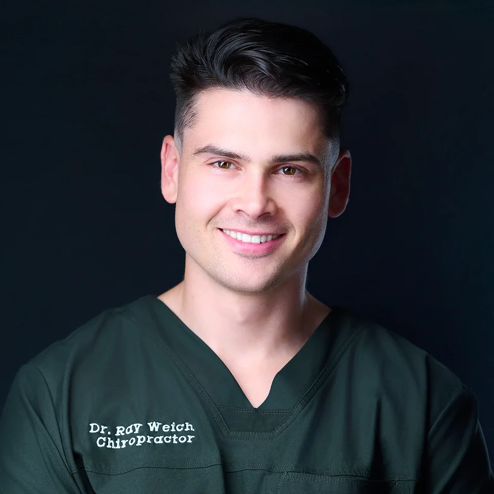 Portrait of a smiling man wearing a dark green medical scrub top with white embroidered text reading 'Dr. Ray Weich Chiropractor' against a solid dark background.