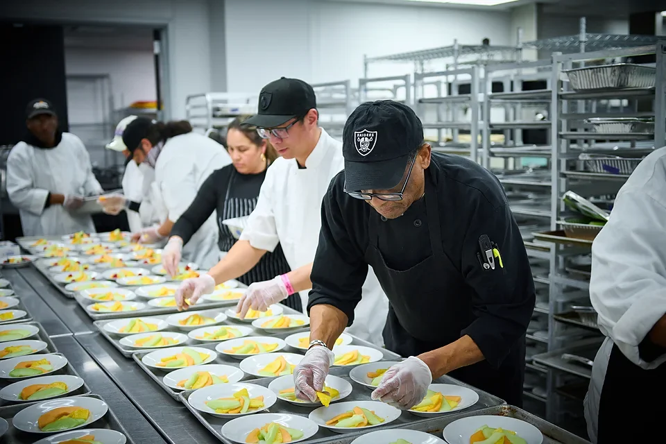 Chefs and kitchen staff preparing multiple plates of fruit desserts in a commercial kitchen with stainless steel shelves.