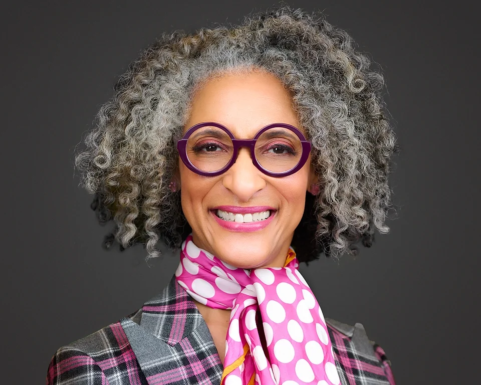Portrait of a woman with curly gray hair, purple glasses, pink lipstick, pink polka dot scarf, and plaid blazer, smiling against a dark background.