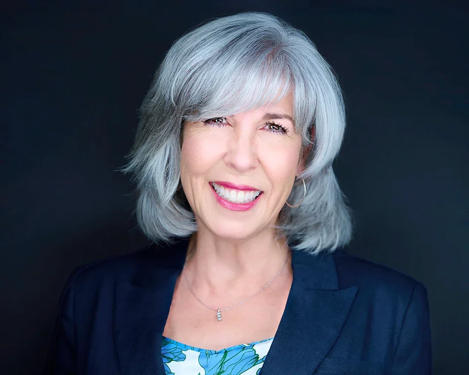 Portrait of a cheerful older woman with gray hair wearing a dark blazer and a floral top, smiling against a dark background.