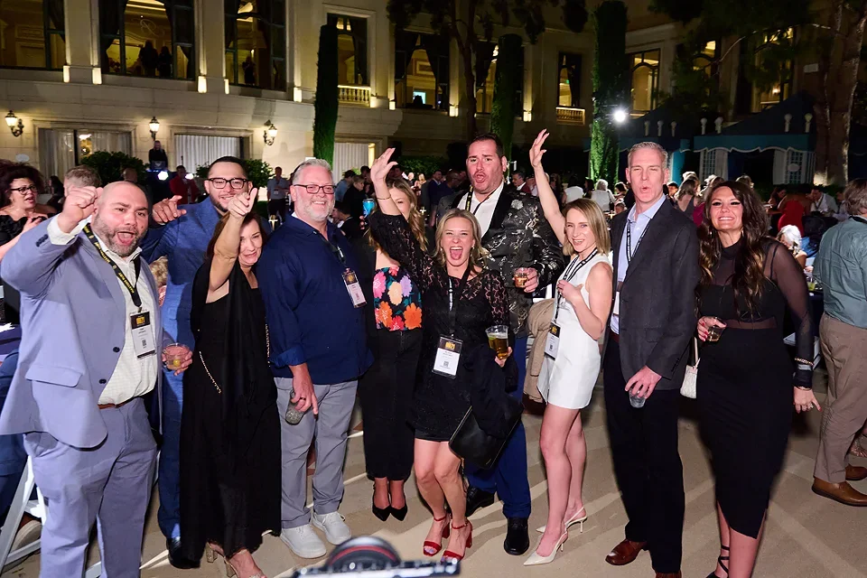 A group of nine diverse people celebrating at a formal event, standing close together and smiling, some holding drinks, in a decorated indoor venue with high ceilings and lighting.