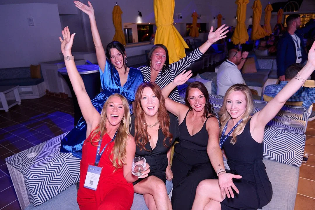A group of five women celebrating at a party, sitting on a couch with their arms raised, smiling, and dressed in colorful and elegant outfits, with a background of yellow umbrellas and other party guests.