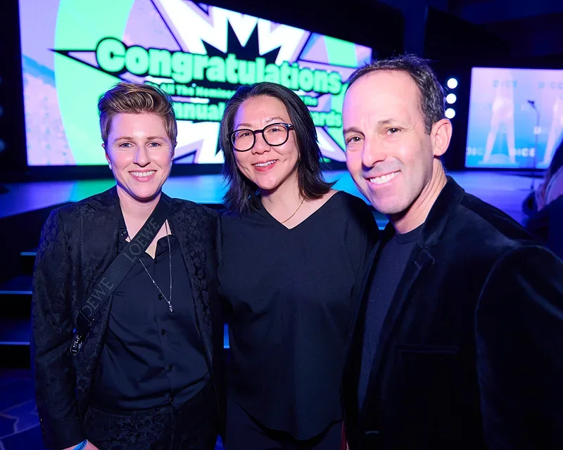 Three people smiling at an awards ceremony, standing in front of a large screen displaying the words 'Congratulations' and 'Awards'.